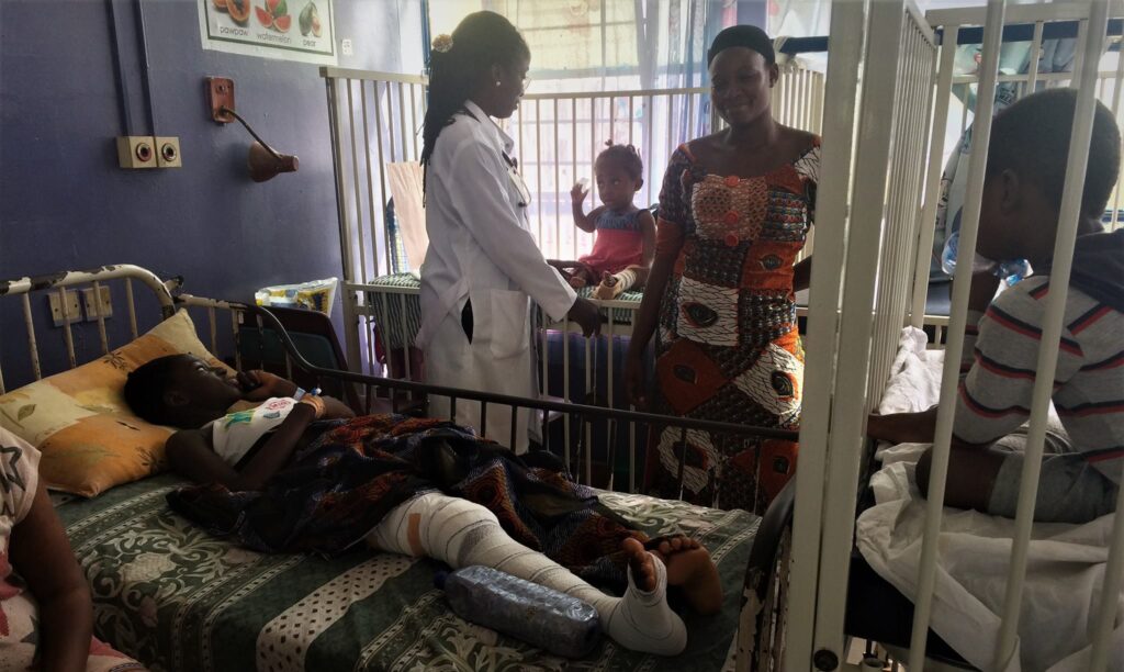 hospital room in Abena Ghana with hospital staff in white coat talking to a lady in a colorful dress, while one child lays in a bed with a bandaged leg and another child and an infant sit in cribs