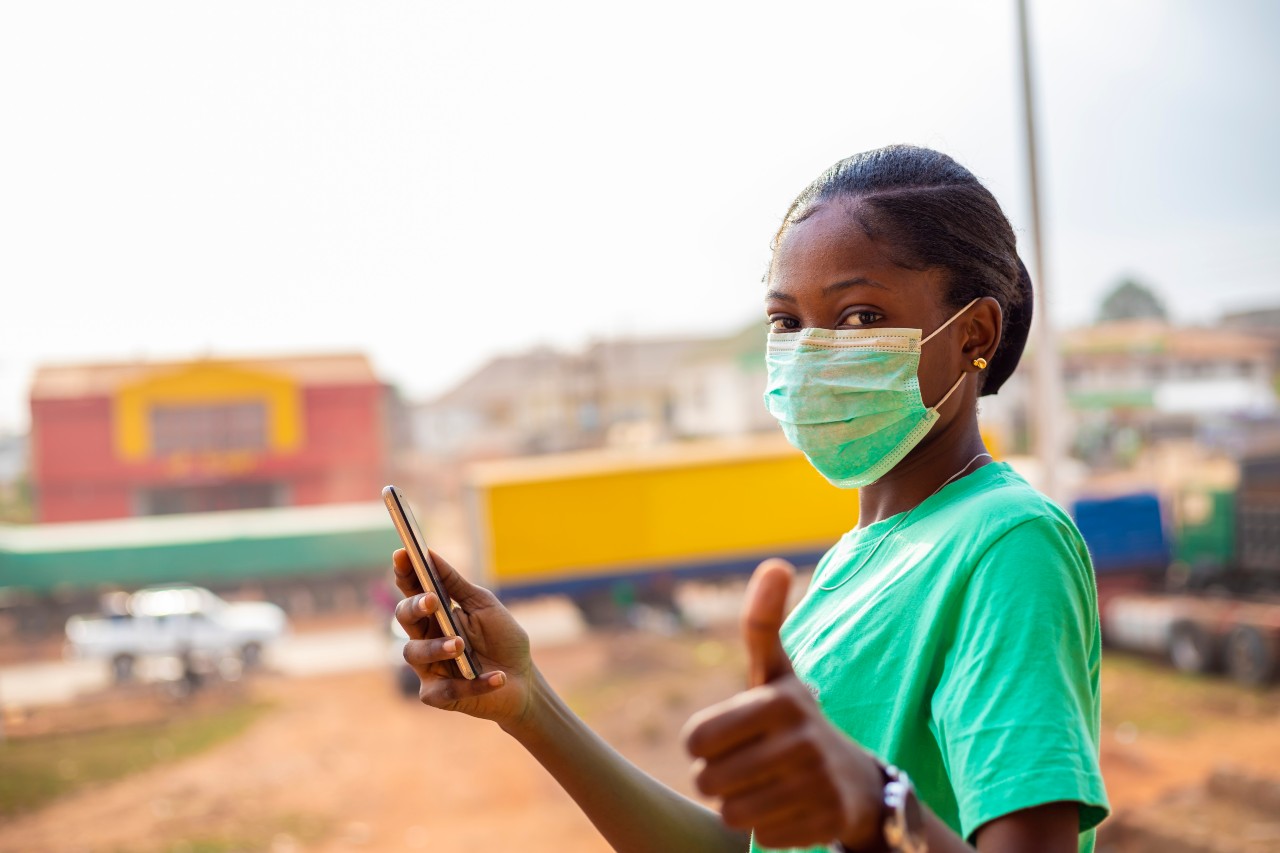 nurse in green scrubs with facenask holding a cellphone and giving a thumbs up sign