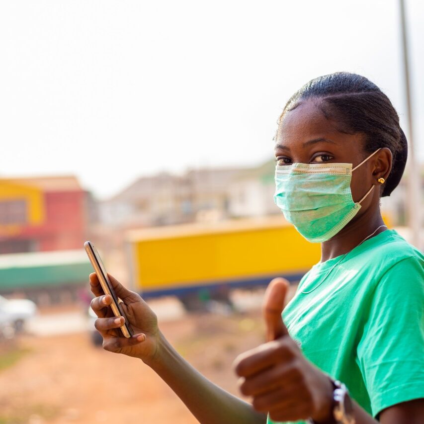 nurse in green scrubs with facenask holding a cellphone and giving a thumbs up sign